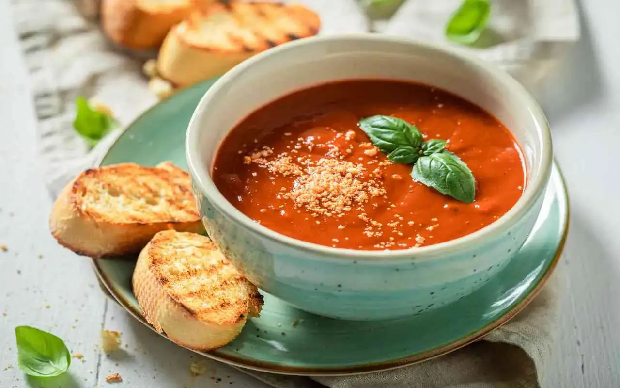Una ciotola di cremosa zuppa di pomodoro, guarnita con basilico fresco e parmigiano, servita con crostini tostati.