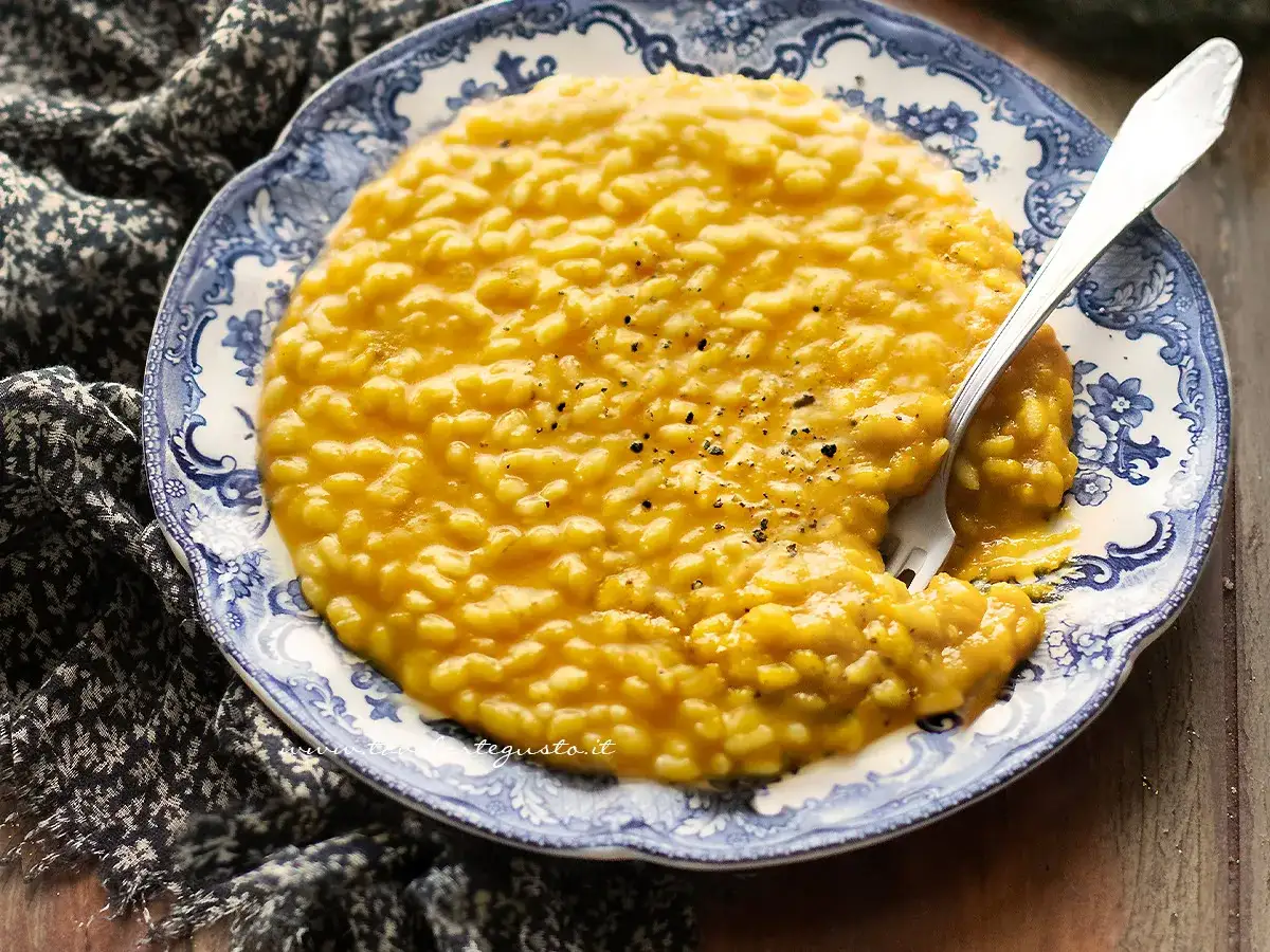 Un cremoso risotto con la zucca, servito su un piatto decorato con motivi blu e bianchi. Una forchetta &egrave; immersa nel piatto, pronta per gustare questo piatto autunnale.