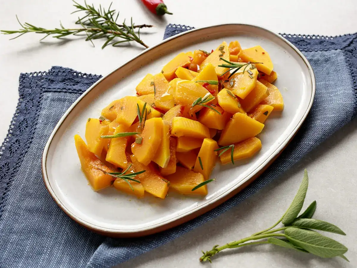 Zucca in padella, tagliata a pezzi e condita con erbe aromatiche, servita su un piatto ovale.