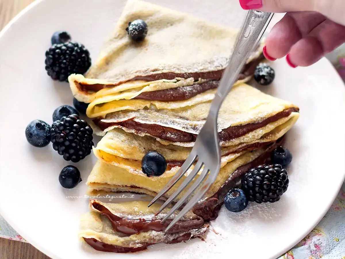Deliziose crepes alla nutella, impilate e guarnite con frutti di bosco freschi e una spolverata di zucchero a velo. Una forchetta &egrave; pronta per assaporare questo dolce.