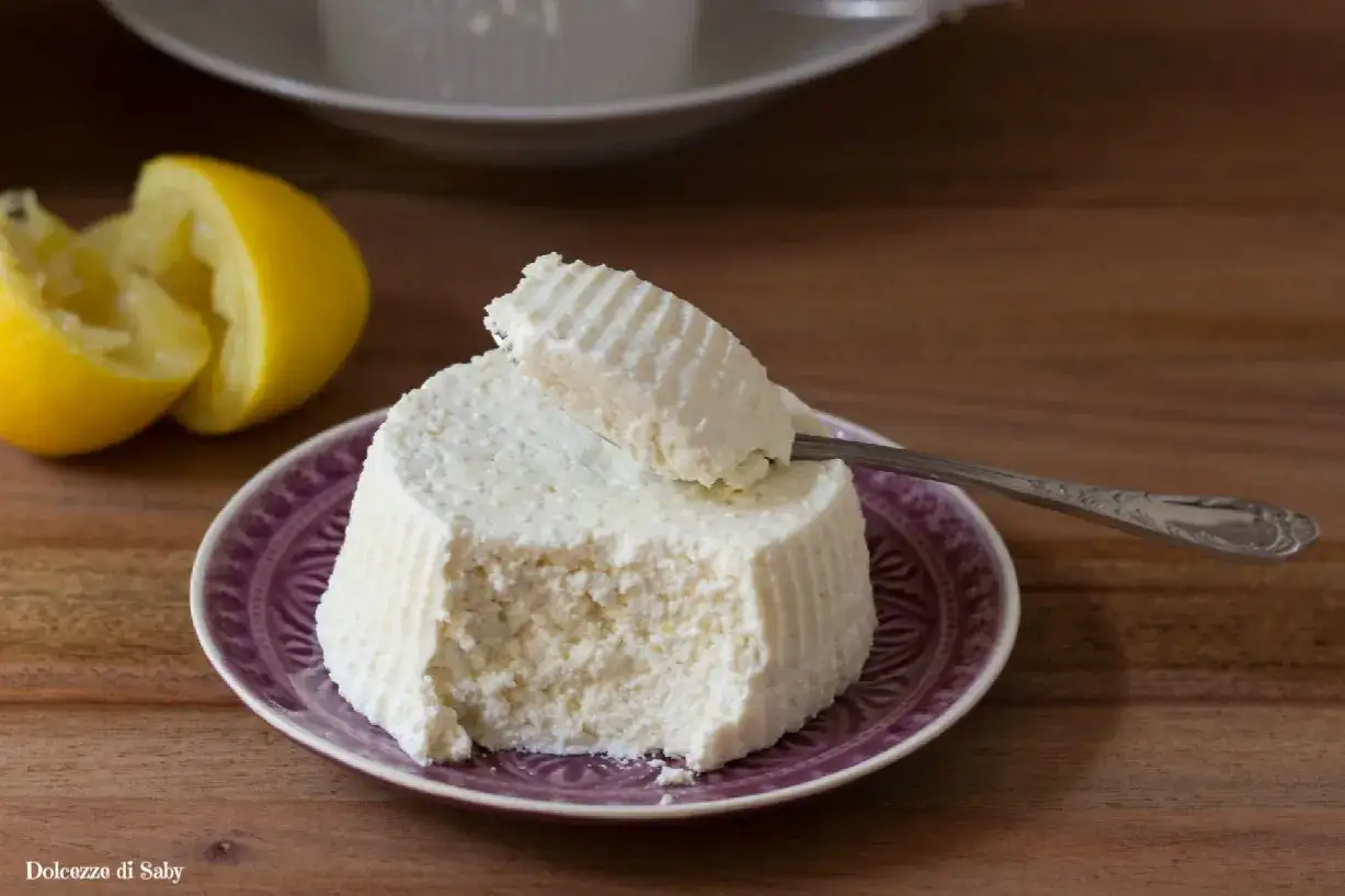 Ricotta fatta in casa su un piatto viola, con limone spremuto accanto. Un cucchiaino preleva una porzione cremosa.