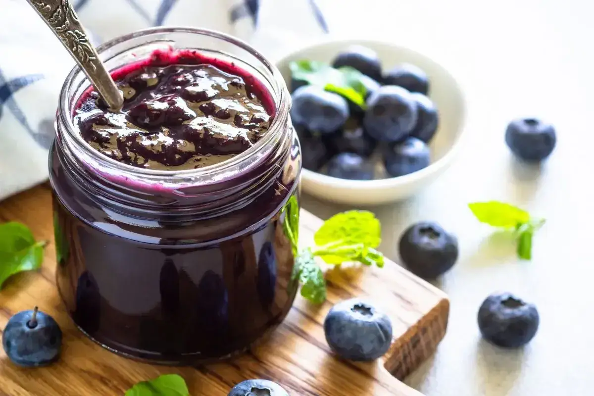 Un vasetto pieno di deliziosa marmellata di mirtilli, con mirtilli freschi e foglie di menta sparse su un tagliere di legno.