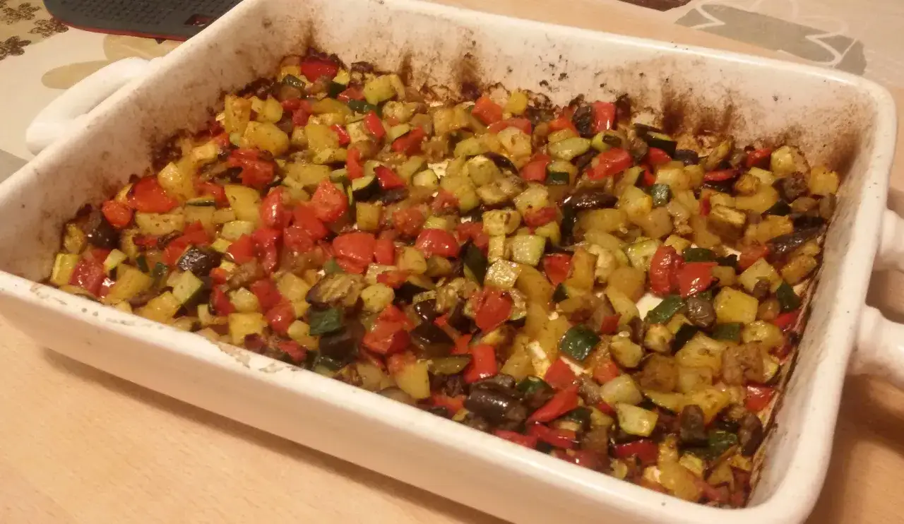 Un tegame bianco pieno di contorni di verdure miste, tra cui peperoni rossi, zucchine e melanzane, appena sfornato e profumato.