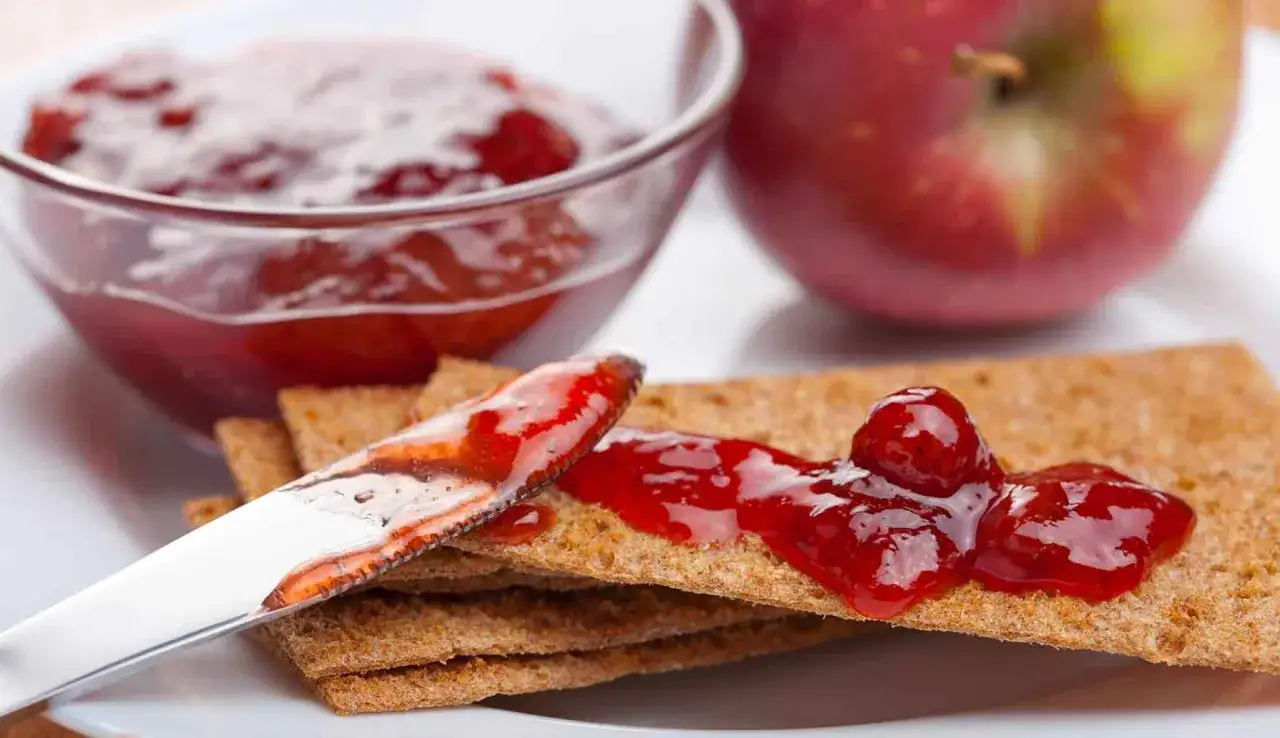 Un coltello spalma marmellata di mele su cracker croccanti, con una ciotola piena e una mela rossa sullo sfondo.