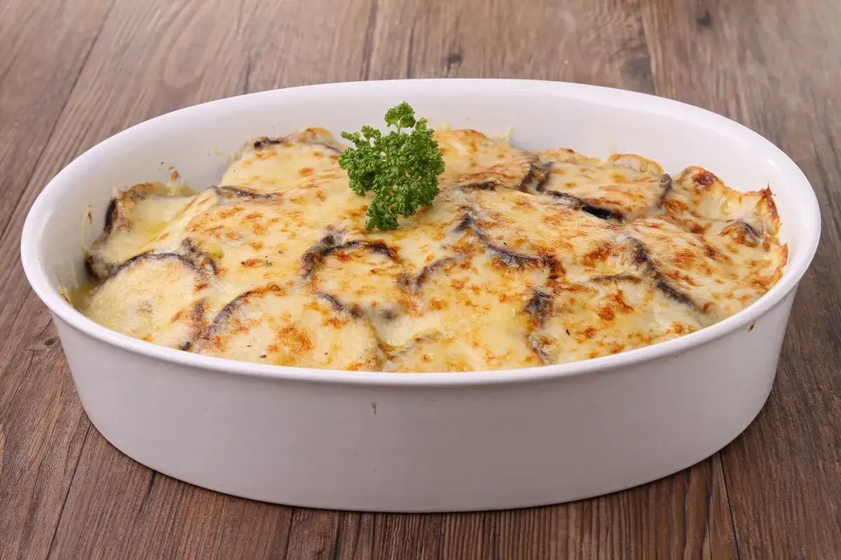 Parmigiana di melanzane bianca, dorata e filante, guarnita con prezzemolo fresco, in una teglia bianca su un tavolo di legno.