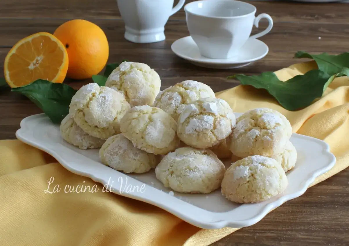 Biscotti con farina di mandorle, spolverati di zucchero a velo, su un piatto bianco, con arance e tazze sullo sfondo.