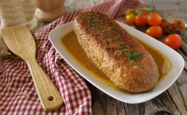 Polpettone in padella dorato e succulento, guarnito con erba cipollina, su un piatto bianco con salsa. Accanto, una spatola di legno e pomodorini.