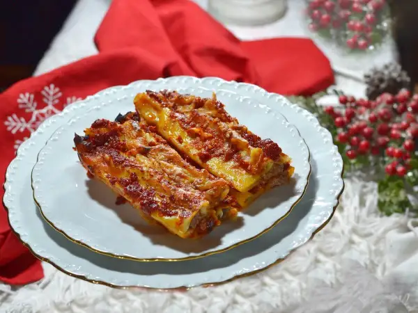 Due cannelloni di carne fumanti, ricoperti di sugo rosso e formaggio fuso, serviti su un piatto bianco con decorazioni natalizie sullo sfondo.