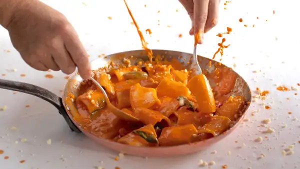 Mani che mescolano paccheri alla Vittorio in padella, con salsa arancione che schizza.