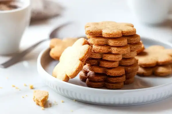 Frolla per biscotti a forma di fiore, impilata su un piatto bianco, con una tazza di caffè sullo sfondo.
