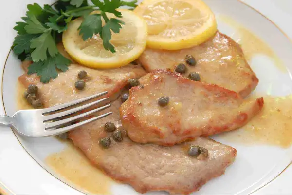 Scaloppine al limone succulente, guarnite con capperi, prezzemolo fresco e fette di limone. Una forchetta attende il primo assaggio.