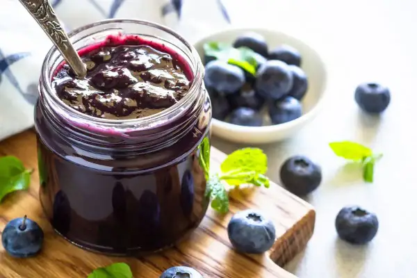 Vasetto di marmellata di mirtilli fatto in casa, con mirtilli freschi e foglie di menta su un tagliere di legno.