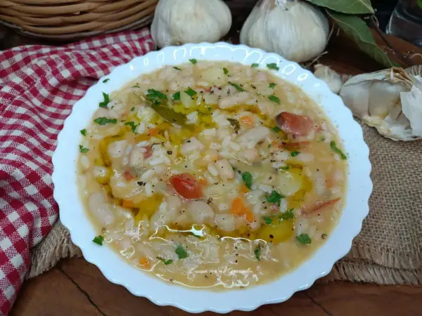 Un piatto fumante di riso e fagioli, condito con olio d'oliva e prezzemolo, con aglio e una tovaglia a scacchi sullo sfondo.