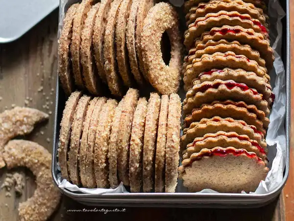 Biscotti grano saraceno ripieni di marmellata, impilati in una scatola di latta. Alcuni biscotti a forma di ciambella e a mezzaluna sono sparsi sul tavolo.