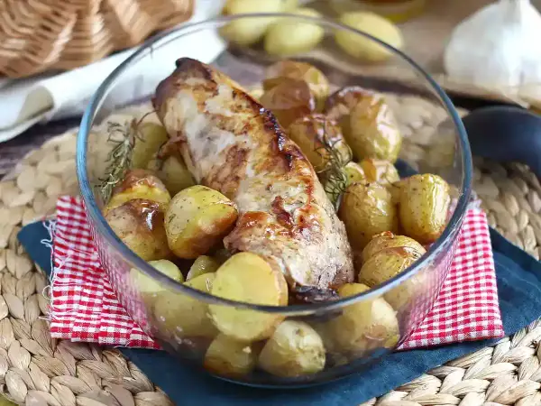 Un succulento filetto di maiale al forno, dorato e profumato, circondato da patate novelle arrosto e rametti di rosmarino in una ciotola di vetro.