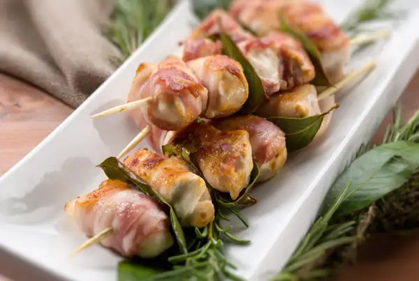 Involtini di lonza avvolti in pancetta, infilzati su spiedini con foglie di alloro e rosmarino.