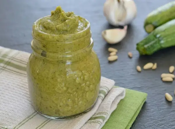 Un vasetto pieno di cremoso pesto di zucchine, con ingredienti freschi come zucchine, aglio e pinoli sullo sfondo.