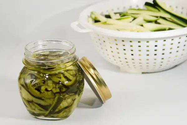 Barattolo di zucchine sott'olio, un modo gustoso per conservare le zucchine. In background, zucchine tagliate a julienne in uno scolapasta.