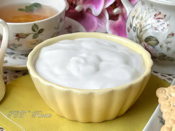 Una ciotola gialla piena di soffice crema al latte, accanto a una tazza di tè e fiori.