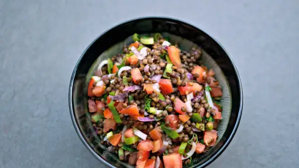 Una fresca insalata di lenticchie estiva, con pomodori, cipolla rossa e erbe aromatiche, servita in una ciotola scura.
